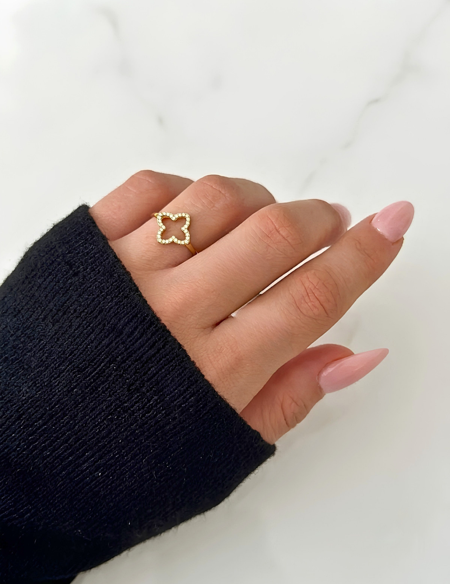 HAND WEARING CLOVER GOLD PLATED RING AND PINK LONG NAILS ON WHITE SURFACE